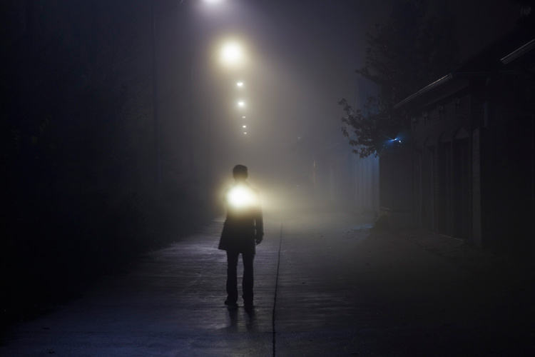 Man searching with flashlight in an alley on a foggy night.