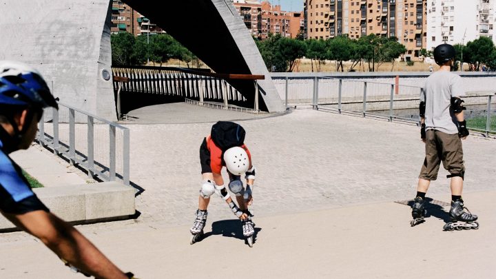 picture of persons riding roller skates in a park beside buildings wearing protective gear
