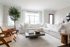 A bright living room with a large white sectional sofa, wooden chairs, and a ceiling fan and light providing soft illumination above.